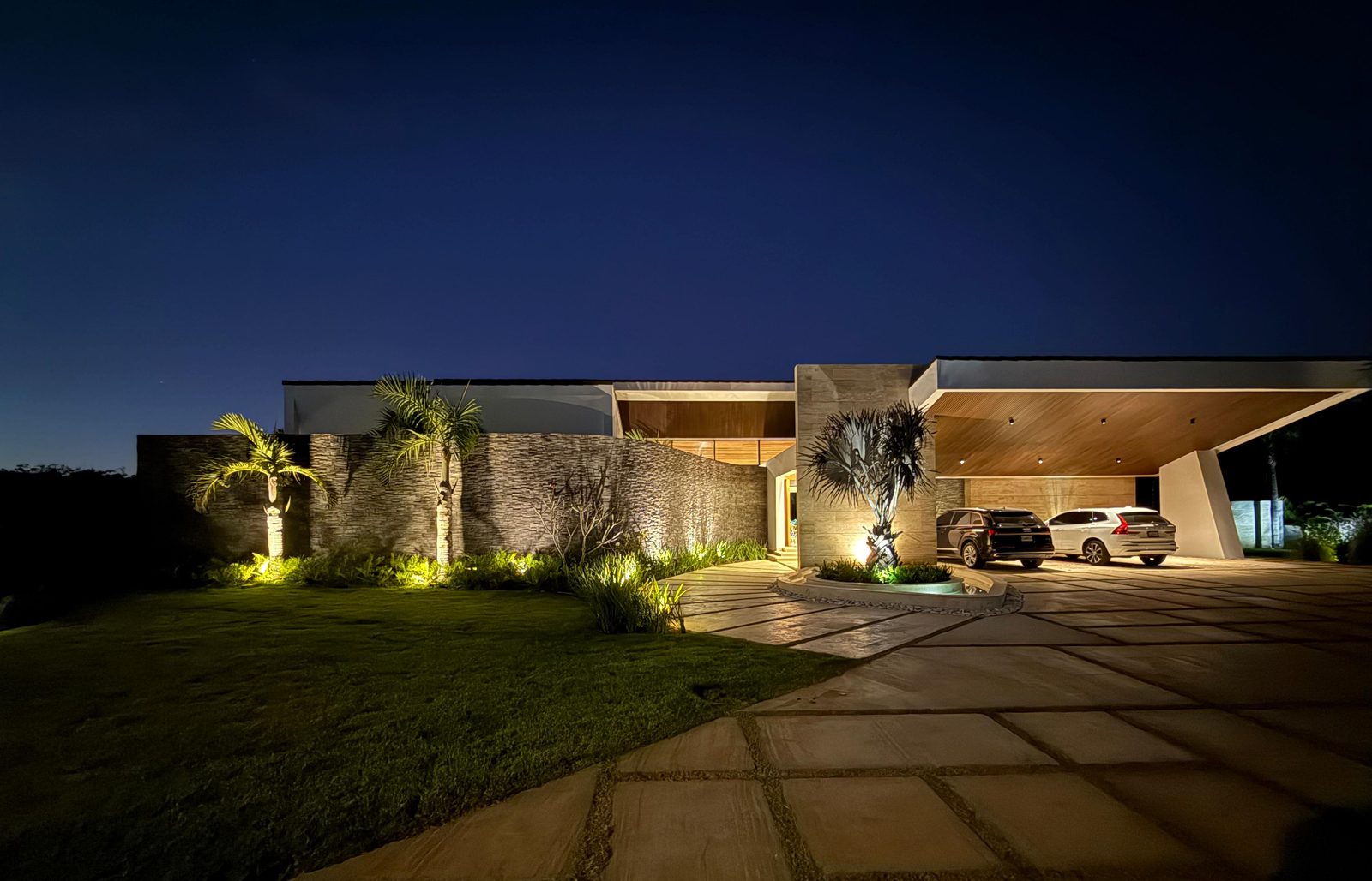 Villa dominicana contemporánea al atardecer, con muro de piedra, palmeras iluminadas y acceso techado para vehículos